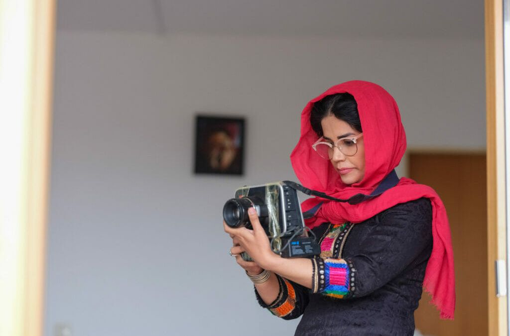 Eine junge Frau trägt ein rotes Kopftuch, unter dem dunkle lockige Haare hervorlugen, dazu ein traditionell afghanisches Gewand mit schwarzer Grundfarbe und rot-pink-violetten Verzierungen an den Ärmeln. Sie ist im Profil zu sehen und schaut konzentriert auf die Kamera, die sie vor sich hält. Im Hintergrund hängt ein kleines Bild an der Wand, das nur unscharf zu sehen ist. Zainab Entezar zählt zu den ersten Afghan:innen, die über das Bundesaufnahmeprogramm nach Deutschland evakuiert wurden.