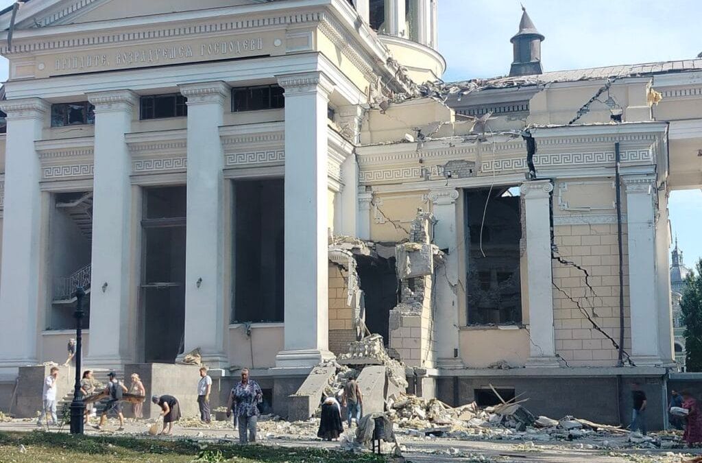 People gather around a heavily damaged classical-style building with debris strewn around.