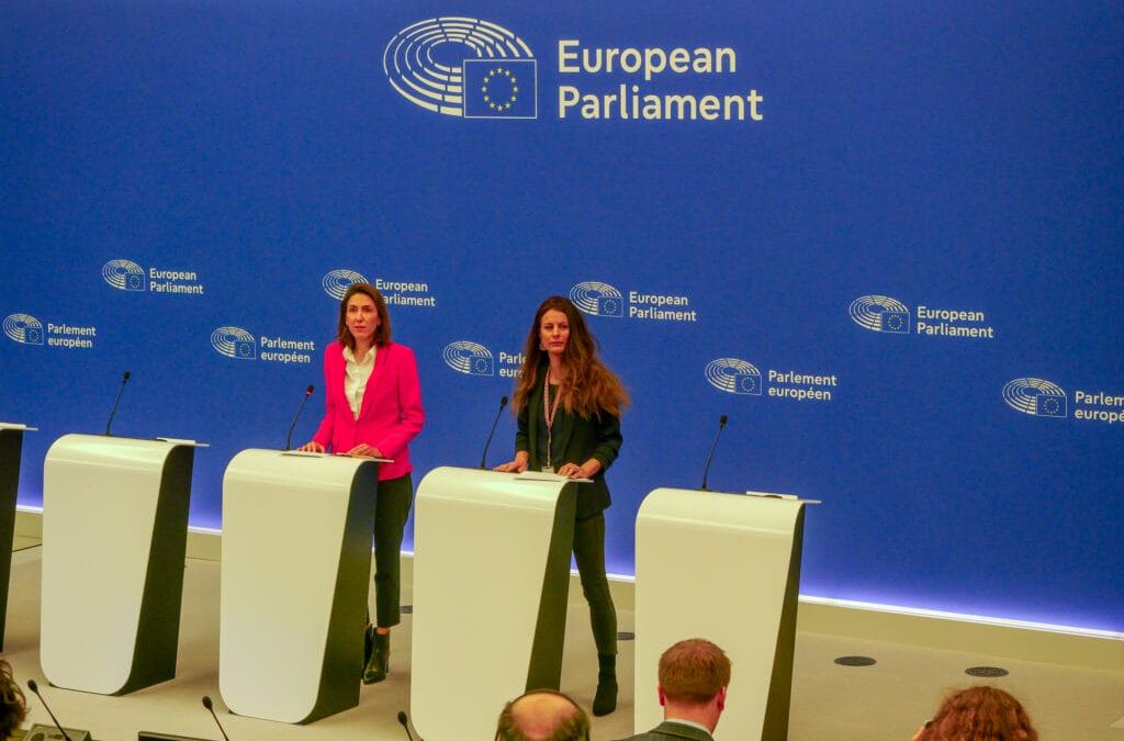 Zwei Frauen stehen an Rednern vor einem blauen EU-Hintergrund und sprechen zu den sitzenden Teilnehmern des Europäischen Parlaments.