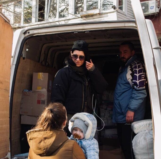 A woman in a black jacket stands inside a van with another man, engaging with a woman holding a warmly clothed baby outside. Various boxes, likely containing food deliveries from Ukrainian volunteers, are visible inside the van.