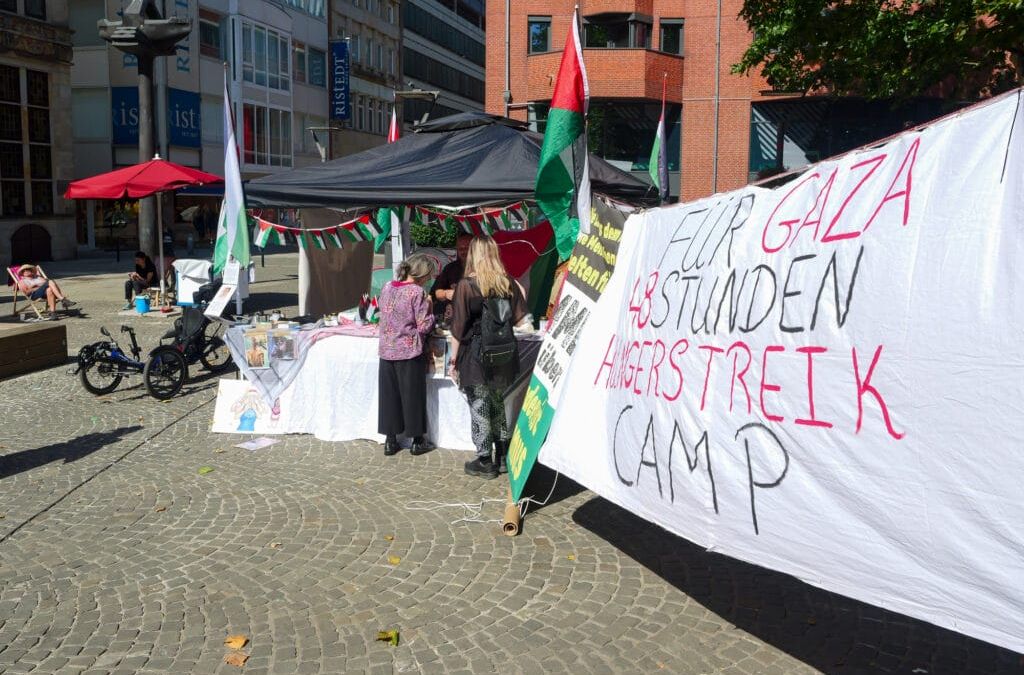 Protestcamp im Freien mit Zelten und Transparenten; auf einem großen Schild steht "Hilf Gaza, 48 Stunden Hungerstreik Camp". Menschen versammeln sich an den Tischen, Palästina- und Gaza-Fahnen sind zu sehen, als David Ginati sich zu den Unterstützern gesellt, um Solidarität zu zeigen.