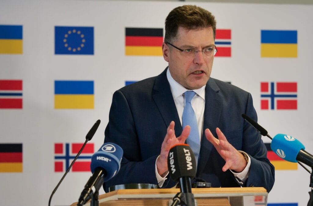Ein Mann spricht an einem Podium mit Mikrofonen und einem Hintergrund mit verschiedenen Nationalflaggen neben der Flagge der Europäischen Union.