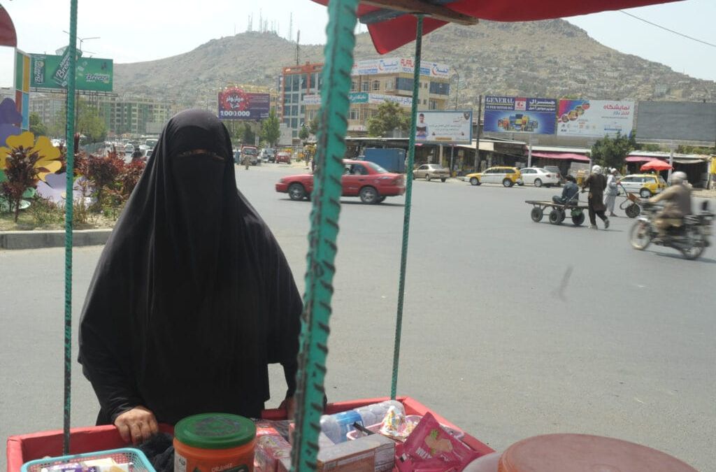 Eine Frau in einem schwarzen Niqab steht an einer belebten Kreuzung in der Stadt an einem mit Waren gefüllten Straßenkarren, im Hintergrund sind Hügel und Gebäude zu sehen.