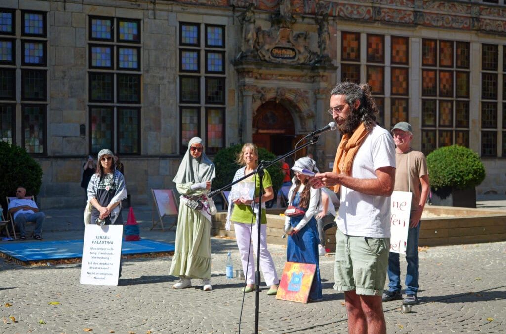 David Ginati spricht bei einer Versammlung im Freien für den Gazastreifen in ein Mikrofon. Hinter ihm stehen mehrere Personen mit Schildern und Plakaten vor einem historischen Gebäude.