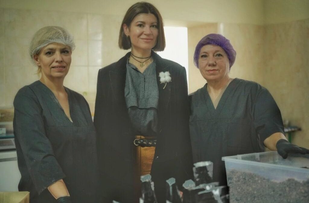 Three women standing together in a workspace: two in protective hairnets and gloves, one in a black outfit with a choker necklace. Surrounded by containers, these Ukrainian volunteers appear dedicated to their mission, possibly preparing to deliver food to those in need, even in the trenches.