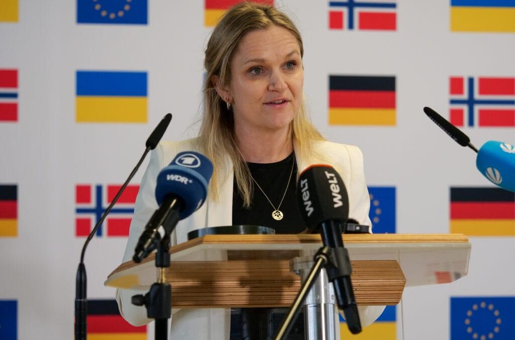 Eine Frau spricht an einem Podium mit Mikrofonen und einem Hintergrund aus EU-Flaggen.