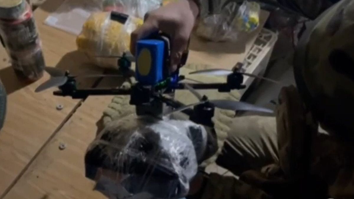 Person in camouflage clothing attaching supplies to a small drone on a wooden surface.