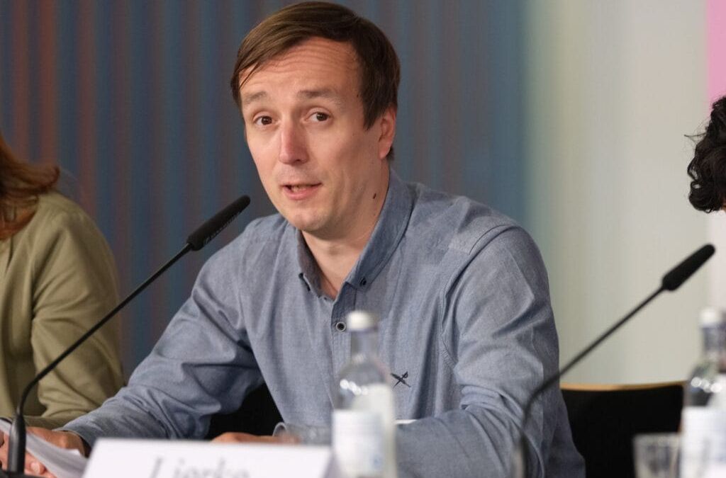 A man in a blue shirt speaks at a conference. In front of him stands a table with microphones and a name badge.