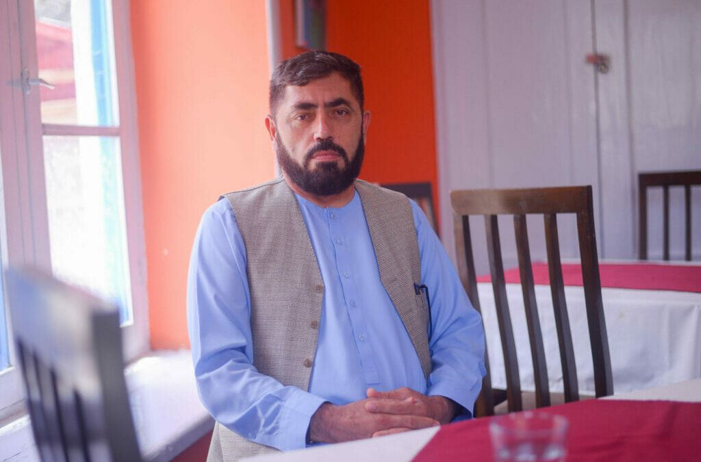 Yasin Adel sitzt mit tradioneller, blauer afghanischer Kleidung und grauer Weste vor einem roten Tisch. Er trägt kurze Haare und einen leichten Vollbart.