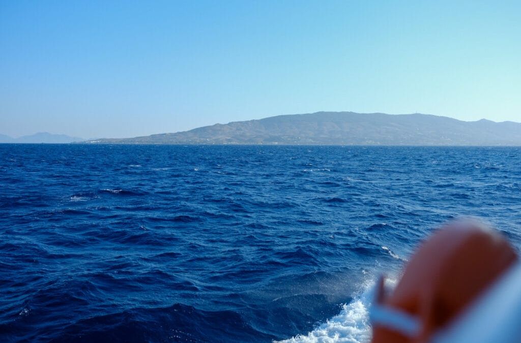 Ein Blick auf das Meer von einem Boot aus, eine Insel türmt sich im Hintergrund auf. Das Meerwasser ist tiefblau und wellig.