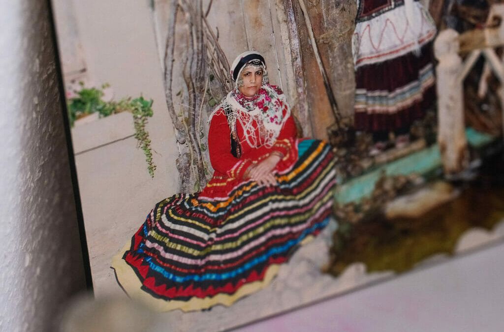 Ein Foto, das fast wie ein Gemälde wirkt: Es zeigt eine Frau in traditioneller Kleidung, die am Boden sitzt, ihr Oberteil ist Rot, der Rock ausgebreitet mit vielen Farbringen.