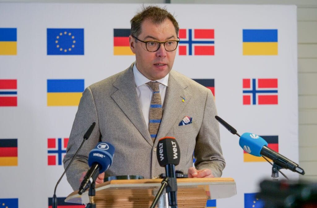 Ein Mann im Anzug spricht auf einem Podium mit Mikrofonen und Flaggen europäischer Länder im Hintergrund.