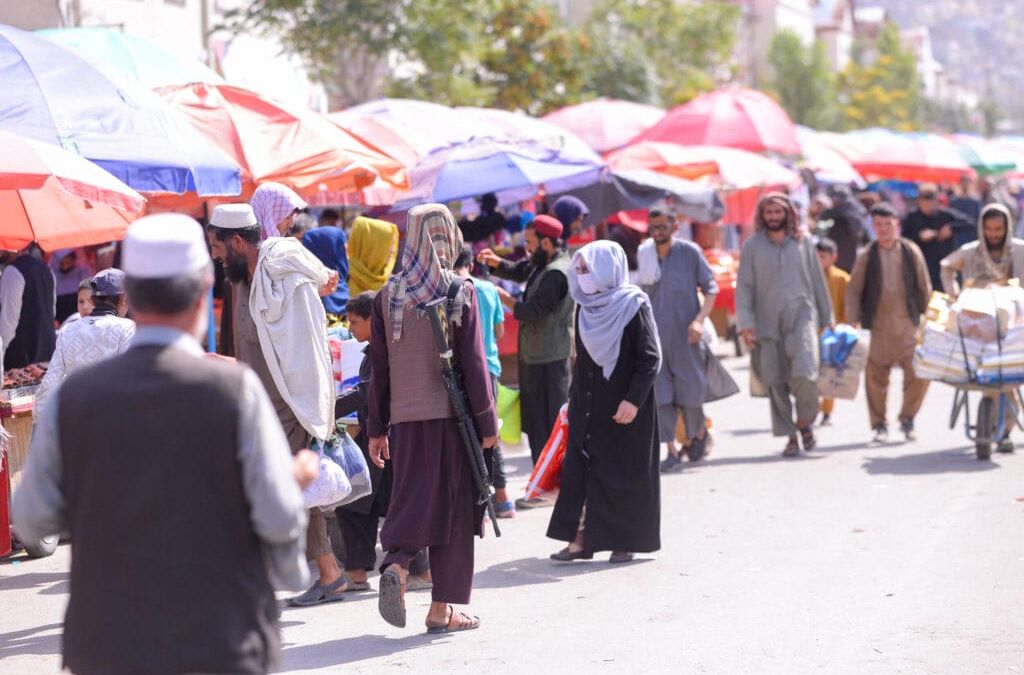 Ein überfüllter Markt im Freien in Kabul, Afghanistan, mit bunten Regenschirmen und Passanten. Eine Person trägt eine Waffe, eine andere schiebt einen mit Kisten beladenen Karren.