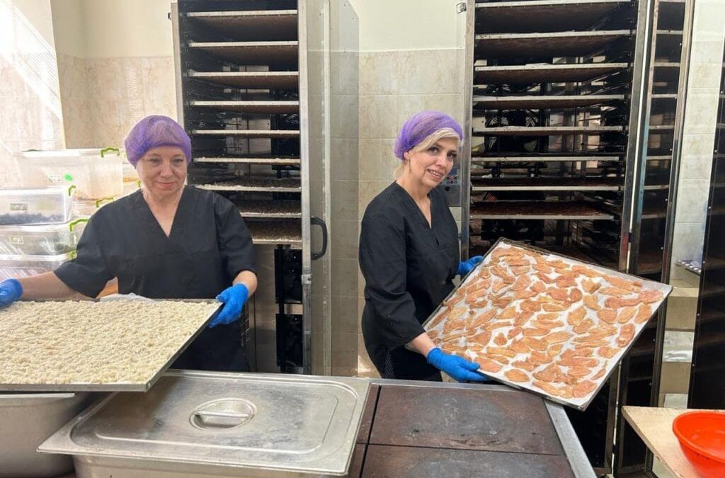 Two Ukrainian volunteers wearing black uniforms, blue gloves, and purple hair nets hold trays of food in a commercial kitchen with multiple ovens, preparing meals for food delivery to the trenches.